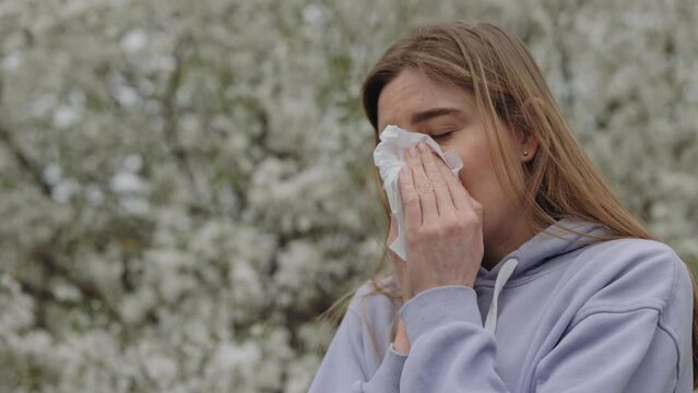 Unhealthy woman blowing nose because of spring pollen allergy outdoors. Side view of sneezing young blonde with napkin in hands among blooming trees in spring park. Health care