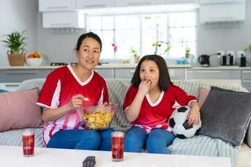 Poster Asian mother and daughter in football shirts watching soccer game, supporting and eating crisps © wavebreak3