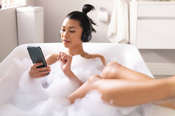 Image of biracial woman relaxing with headphones and smartphone in bathtub in bubble bath