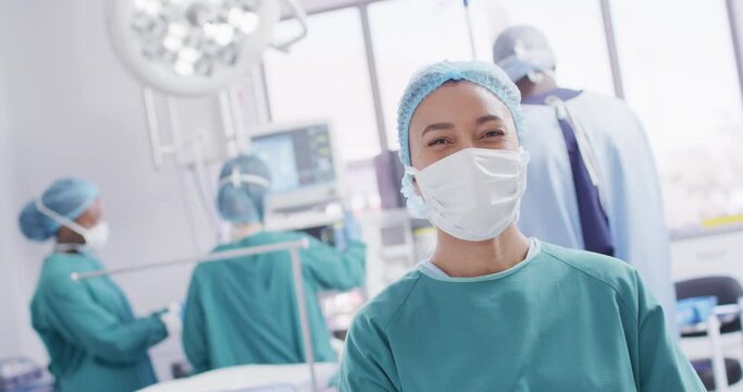 Video portrait of biracial female surgeon in mask smiling in operating theatre, copy space