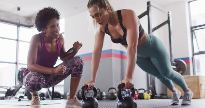 Video of diverse female fitness trainer encouraging woman doing push ups on kettlebells at gym