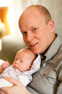 A father holds his newborn smiling baby in his arms in his living room.