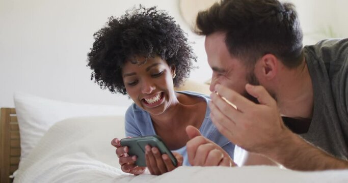 Video of happy diverse couple relaxing at home, lying on bed using smartphone and laughing