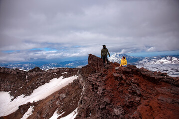 Crater of volcano. There're two people on the edge of crater of Gorely volcano. The man is standing and the woman is sitting. There're red rocks on surface. There's Mutnovskiy volcano in the backgroun