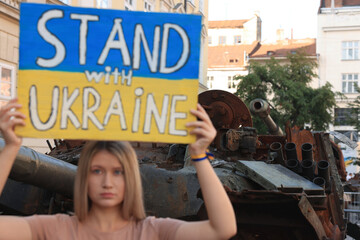 Sad woman holding poster in colors of national flag with words Stand with Ukraine near broken tank on city street