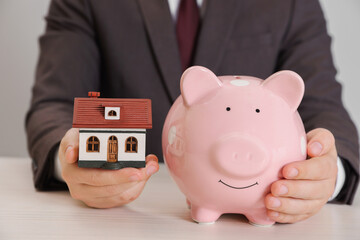 Man holding house model and piggy bank at wooden table, closeup. Saving money concept