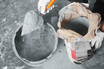 Worker with cement powder and trowel mixing concrete in bucket indoors, closeup