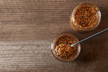 Jars and spoon of whole grain mustard on wooden table, flat lay. Space for text
