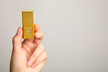 Woman holding shiny gold bar on light grey background, closeup. Space for text