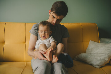 Father dresses infant in living room sitting on couch at home. Clothes baby undershirts and sliders on baby close-up. Home care and baby care concept. - obrazy, fototapety, plakaty