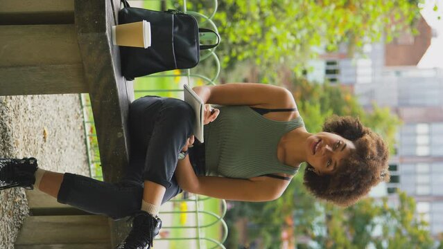 Vertical video of businesswoman sitting on bench in city park writing in notebook - shot in slow motion