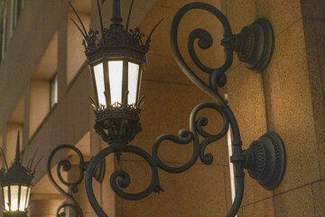 Fototapeta na wymiar Lit Wrought Iron Street Lamp With Sharp Spikes Mounted On The Wall At Night