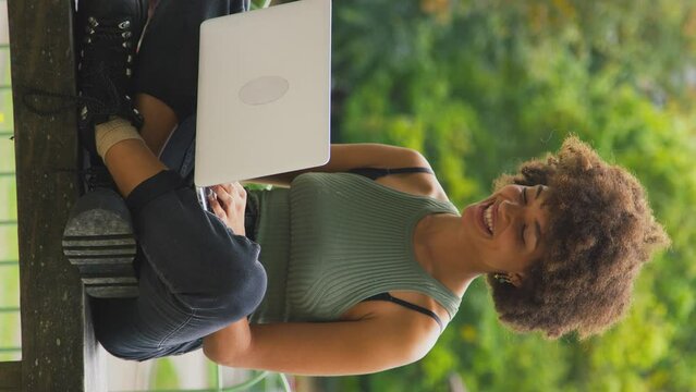 Vertical video of businesswoman sitting on bench in city park working on laptop with takeaway drink - shot in slow motion
