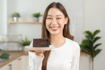 Portrait of attractive pretty girl, asian young teenage woman holding plate of a piece of chocolate cake, enjoy sweet eating dessert meal in kitchen at home. Dieting, diet for loss weight to slim.