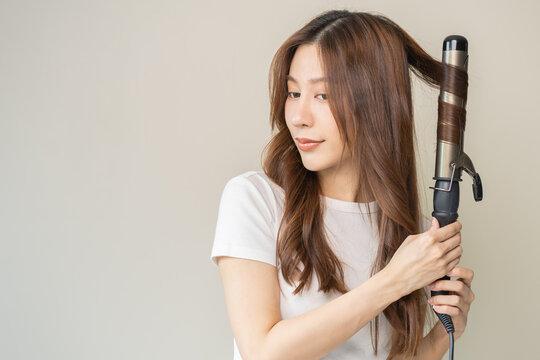 Beautiful curly brunette, attractive asian young woman, girl smiling using curler, curling iron long hair with ceramic curler straightener at home. Hairstyle and Hairdressing. Beauty and hair care.