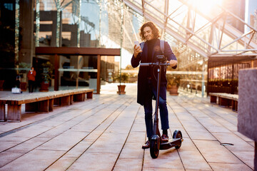 Smiling Hispanic businessman with electric scooter text messaging on cell phone outdoors.