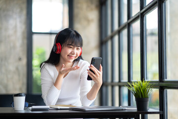 Young adult happy smiling Asian student wearing headphones talking in online chat meetings using a laptop on a university campus or at a virtual office. College female students learning remotely.