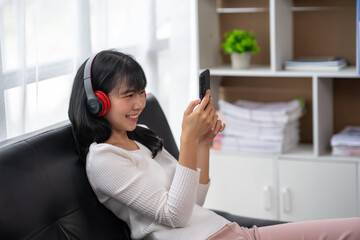 Portrait of a happy young Asian woman listening to music with wireless headphones from a music application on a mobile smartphone in the living room at home