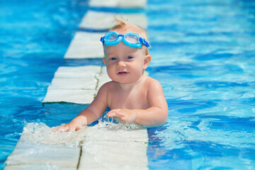 Happy smiling baby with underwater goggles is having fun playing with splashes in blue water in pool before swimming lessons. Lifestyle and summer children activity in vacation in tropical resort