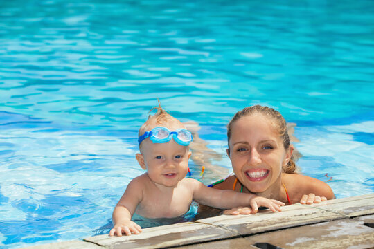 Happy smiling baby in underwater goggles is having fun with young mother in blue water in pool before swimming lessons. Lifestyle and summer children activity in vacation in tropical resort