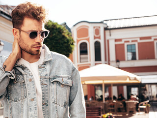 Portrait of handsome confident stylish hipster model. Sexy man dressed in jeans jacket. Fashion male posing in the street. In sunglasses in Europe city. Outdoors at sunset