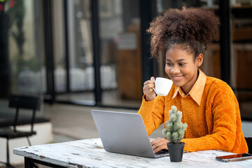 Attractive african young confident woman holding coffee cup and using laptop computer, sitting at open co-workspace. - obrazy, fototapety, plakaty