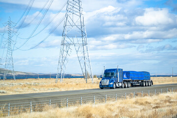 Blue big rig heavy duty semi truck with low profile cab transporting covered and fastened cargo on flat bed semi trailer with additional wheel axles driving on the flat highway road in California - obrazy, fototapety, plakaty