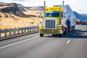 Classic bright yellow powerful big rig semi truck with oversize load sign on the bumper transporting oversized cargo on step down semi trailer driving on the highway road at sun shine