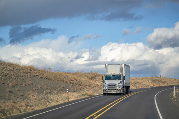 Obraz premium Front view of white big rig semi truck with dry van semi trailer driving downhill on the marked road with clouds on the background