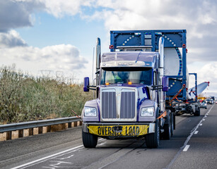Powerful dark blue classic big rig semi truck tractor with oversize load sign on the bumper and additional trolley transporting super long blade from a wind turbine with an escort car