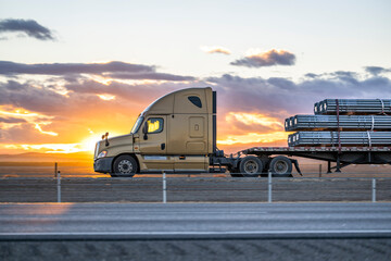Profile of big rig beige semi truck transporting cargo on flat bed semi trailer driving on the one way highway road at sunset time