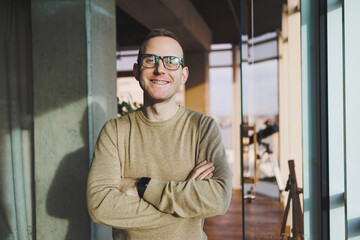 A young man in glasses and casual clothes stands in a modern workspace. Male freelancer works remotely.