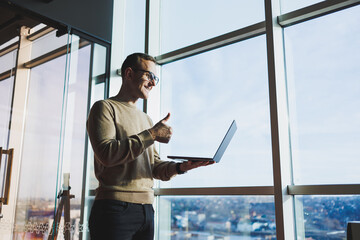 A cute young man in a brown sweater and glasses is standing near a large window in the office with a laptop in his hands while working in the office. A young freelancer works remotely.