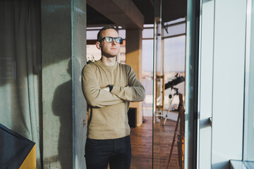 A young man in glasses and casual clothes stands in a modern workspace. Male freelancer works remotely.