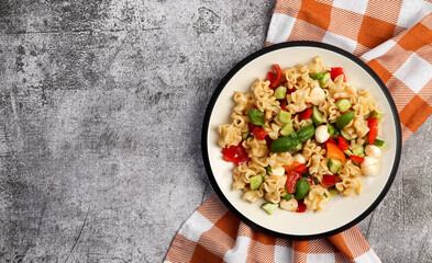 Pasta salad with mozzarella, greens, cucumbers and tomatoes on a round plate on a dark gray...