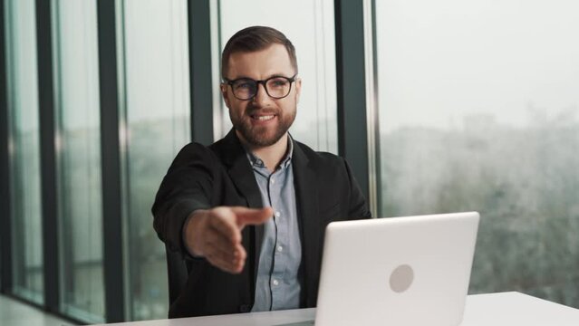 A smiling man in a jacket and glasses shakes hands with an obvious employee of the company in the office sitting at a table with a laptop