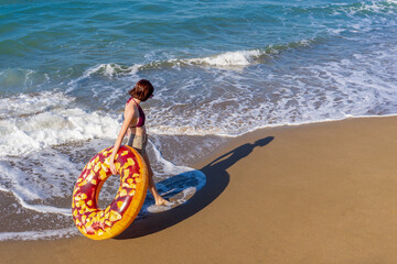 Teen girl with donut lilo walking at sea shore on sand and waves in summer sunny day. Beautiful young woman in swimwear and sunglasses with inflatable ring on beach. Vacation, happy lifestyle concept