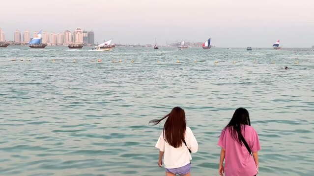 local Qatari boats with world cup countries' flags on it pass. friends spend time on the beach. women fill free and safe.tourists swim and taking photo. Katara beach, Doha, Qatar 2022.