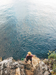 High angle view of unrecognizable male with straw hat fishing with rod at rocky seaside coastline