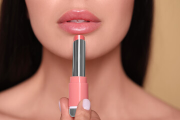 Young woman applying nude lipstick on beige background, closeup