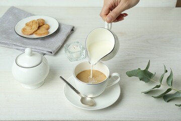 Woman pouring milk into cup with coffee at white wooden table, closeup