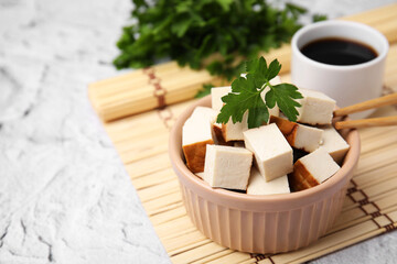 Bowl of smoked tofu cubes, soy sauce and parsley on white textured table, closeup. Space for text