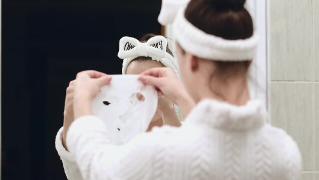 A young woman in a bathrobe and a towel on her head after a morning shower stands in front of a mirror and fixes a moisturizing sheet mask on her face