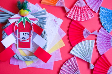 しめ縄とランダムに配置した折り紙の扇と和紙と赤背景の写真。しめ縄には迎春と書いてある