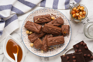 Delicious chocolate candy bars, caramel and nuts on white marble table, flat lay