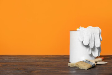 Can of paint, gloves and brushes on wooden table against orange background. Space for text