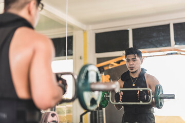 Fototapeta premium A man does hammer curls with a tricep bar. Checking his form in the mirror. Working out near the dumbbell rack near the gym.