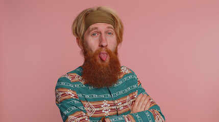 Funny joyful sincere redhead hippie man in pattern shirt making playful silly facial expressions and grimacing, fooling around showing tongue. Young handsome hipster guy isolated on pink background