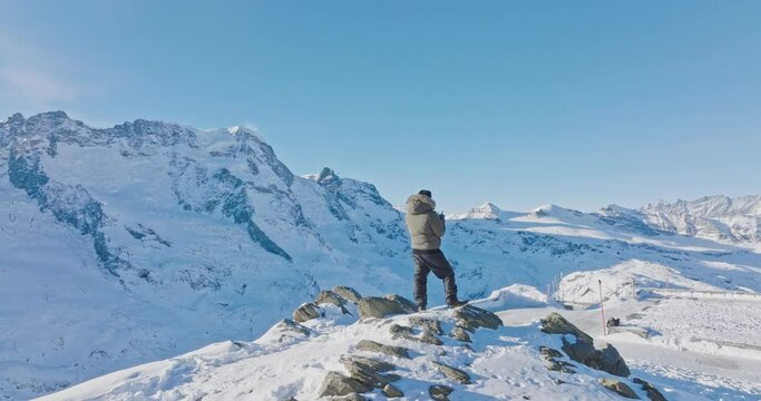 Aerial orbit motion of Young asian man standing on top of the famous Gornergrat observatory operates with drone remote control with iconic famous Matterhorn sunset background. Swiss alps, Switzerland.