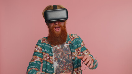 Redhead bearded hippie man using headset helmet app to play simulation innovation game. Watching virtual reality 3D 360 video. Young ginger guy boy in VR goggles isolated on pink studio background